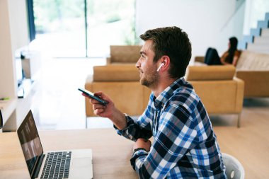 Modern bir oturma odasındaki masada oturan bir serbest çalışan, kulaklıklarıyla iş görüşmesi için bir laptop kullanan, arkadaşlarıyla sohbet eden ve eğlenen biri..
