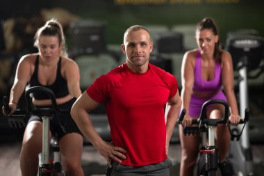 Kararlı bir fitness eğitmeni, iki bayan müşterisinin önünde, arka plandaki antrenmanlarında onlara rehberlik eder.