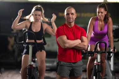 Kararlı bir fitness eğitmeni, iki bayan müşterisinin önünde, arka plandaki antrenmanlarında onlara rehberlik eder.