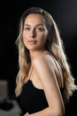 Parlak siyah elbiseli, nefes kesici sarışın bir kadın stüdyoda zarifçe poz veriyor, özgüven, zarafet ve ebedi güzellik yayıyor..