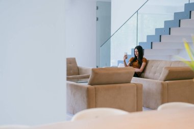 Modern bir evde otururken akıllı telefonu video görüşmesi için kullanan bir kadın.