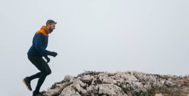 Atletik kıyafetli bir adam sisli havada kayalık bir tepeye tırmanır. Mavi ceket ve siyah tayt giymişti. Soluk, sisli bir arka plana karşı kararlı bir hareketle orta adımda yakalandı.. 