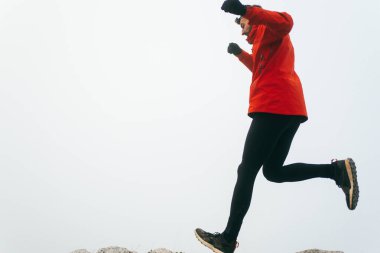 Atletik kıyafetli bir adam sisli havada kayalık bir tepeye tırmanır. Kırmızı ceket ve siyah tayt giymişti. Soluk, sisli bir arka plana karşı kararlı bir hareketle orta adımda yakalandı.. 