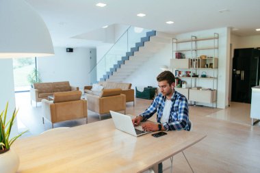 Modern bir oturma odasındaki masada oturan bir serbest çalışan, kulaklıklarıyla iş görüşmesi için bir laptop kullanan, arkadaşlarıyla sohbet eden ve eğlenen biri..