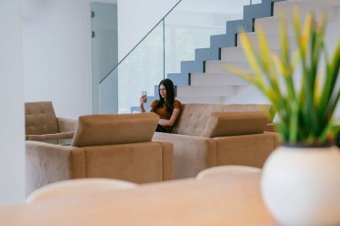 Modern bir evde otururken akıllı telefonu video görüşmesi için kullanan bir kadın.