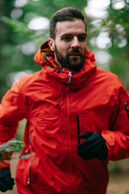 Kırmızı ceket giyen bir adam uzun çam ağaçları ve sonbahar yapraklarıyla çevrili bir orman yolunda koşuyor. Sakin orman yolu, güz mevsiminde doğada barışçıl bir doğa aktivitesi olduğunu gösteriyor.