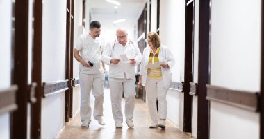 Hasta belgelerini inceleyip tedavi planlarını tartışırken beyaz üniformalı bir grup sağlık görevlisi hastane koridorunda yürüyorlar. 