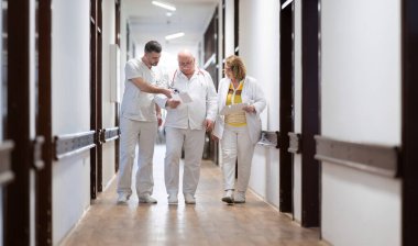 Hasta belgelerini inceleyip tedavi planlarını tartışırken beyaz üniformalı bir grup sağlık görevlisi hastane koridorunda yürüyorlar. 