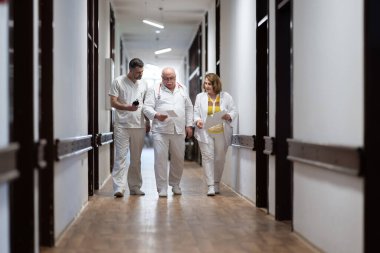 Hasta belgelerini inceleyip tedavi planlarını tartışırken beyaz üniformalı bir grup sağlık görevlisi hastane koridorunda yürüyorlar. 