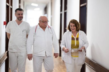 Hasta belgelerini inceleyip tedavi planlarını tartışırken beyaz üniformalı bir grup sağlık görevlisi hastane koridorunda yürüyorlar. 