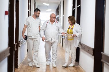 Hasta belgelerini inceleyip tedavi planlarını tartışırken beyaz üniformalı bir grup sağlık görevlisi hastane koridorunda yürüyorlar. 