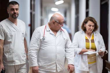 Hasta belgelerini inceleyip tedavi planlarını tartışırken beyaz üniformalı bir grup sağlık görevlisi hastane koridorunda yürüyorlar. 