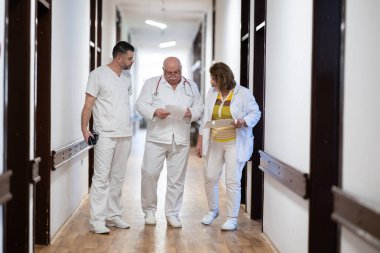Hasta belgelerini inceleyip tedavi planlarını tartışırken beyaz üniformalı bir grup sağlık görevlisi hastane koridorunda yürüyorlar. 