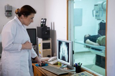 Beyaz laboratuvar önlüklü bir radyolog, bir hastanın kalça röntgenini incelerken bir çalışma odasında oturuyor.