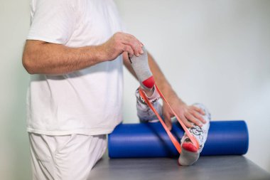 Terapist, fizik tedavi seansında direnç bandı kullanarak bacak esnetmeye yardım ediyor..