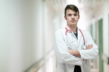 Kollarını kavuşturup hastane koridorunda duran beyaz önlüklü genç bir doktorun portresi..