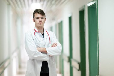 Kollarını kavuşturup hastane koridorunda duran beyaz önlüklü genç bir doktorun portresi..