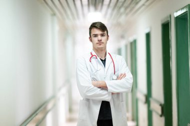 Kollarını kavuşturup hastane koridorunda duran beyaz önlüklü genç bir doktorun portresi..