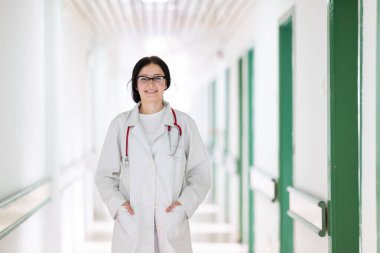 Kendine güvenen, steteskoplu genç bir bayan doktor. Parlak bir hastane koridorunda dururken gülümsüyor.. 