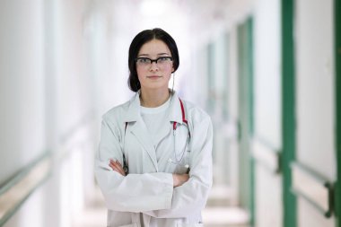 Kendine güvenen, steteskoplu genç bir bayan doktor. Parlak bir hastane koridorunda dururken gülümsüyor.. 