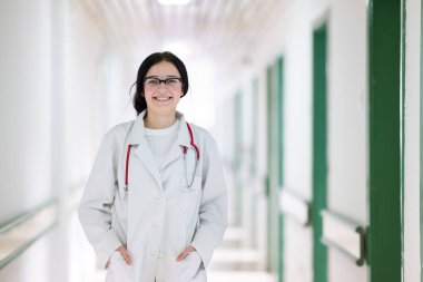 Kendine güvenen, steteskoplu genç bir bayan doktor. Parlak bir hastane koridorunda dururken gülümsüyor.. 