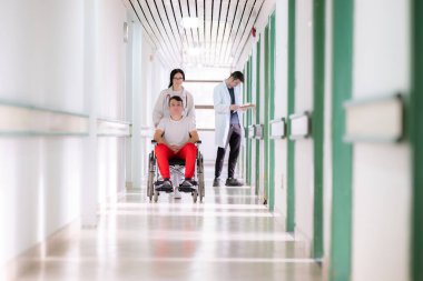 Tekerlekli sandalyedeki gülümseyen genç bir adam beyaz önlüklü ve steteskoplu bir kadın sağlık görevlisi tarafından itiliyor. Hastane koridorundalar, tıbbi ya da rehabilitasyon ortamı öneriyorlar.