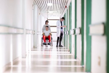 Tekerlekli sandalyedeki gülümseyen genç bir adam beyaz önlüklü ve steteskoplu bir kadın sağlık görevlisi tarafından itiliyor. Hastane koridorundalar, tıbbi ya da rehabilitasyon ortamı öneriyorlar.