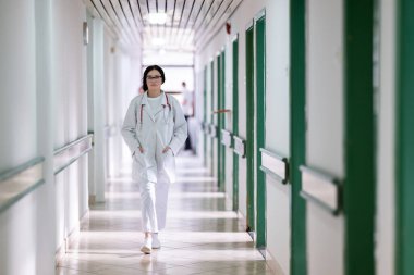 Kendine güvenen, steteskoplu genç bir bayan doktor. Parlak bir hastane koridorunda dururken gülümsüyor.. 