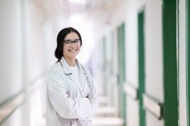 Kendine güvenen, steteskoplu genç bir bayan doktor. Parlak bir hastane koridorunda dururken gülümsüyor.. 