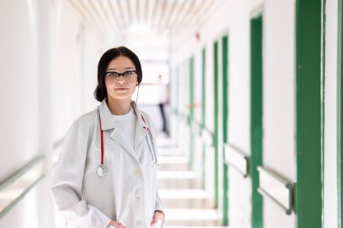 Kendine güvenen, steteskoplu genç bir bayan doktor. Parlak bir hastane koridorunda dururken gülümsüyor.. 