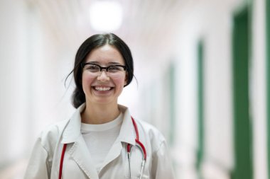 Kendine güvenen, steteskoplu genç bir bayan doktor. Parlak bir hastane koridorunda dururken gülümsüyor.. 