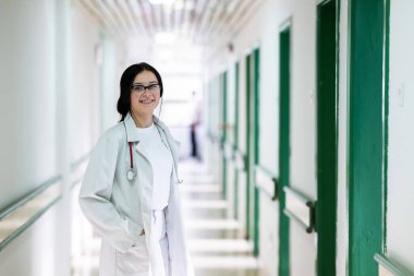 Kendine güvenen, steteskoplu genç bir bayan doktor. Parlak bir hastane koridorunda dururken gülümsüyor.. 
