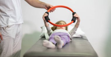 Genç bir kız, bir antrenörün yardımıyla rehabilitasyon egzersizleri yapıyor. Tıp kürsüsünde yatarken direniş halkası kullanıyor..