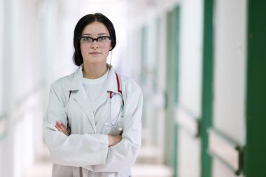 Kendine güvenen, steteskoplu genç bir bayan doktor. Parlak bir hastane koridorunda dururken gülümsüyor.. 