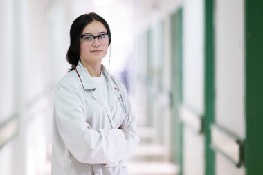 Kendine güvenen, steteskoplu genç bir bayan doktor. Parlak bir hastane koridorunda dururken gülümsüyor.. 