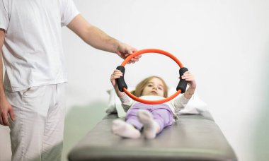 Genç bir kız, bir antrenörün yardımıyla rehabilitasyon egzersizleri yapıyor. Tıp kürsüsünde yatarken direniş halkası kullanıyor..