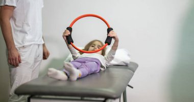 Genç bir kız, bir antrenörün yardımıyla rehabilitasyon egzersizleri yapıyor. Tıp kürsüsünde yatarken direniş halkası kullanıyor..