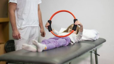 Genç bir kız, bir antrenörün yardımıyla rehabilitasyon egzersizleri yapıyor. Tıp kürsüsünde yatarken direniş halkası kullanıyor..