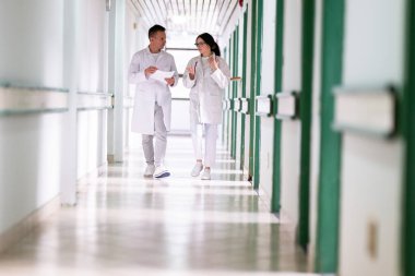 Beyaz önlüklü iki doktor, hastane koridorunda yürüyüp tıbbi belgeleri tartışıyorlar. Takım çalışmalarını ve profesyonel sağlık hizmetlerini ön plana çıkarıyorlar.. 