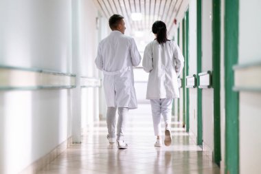 Beyaz önlüklü iki doktor, hastane koridorunda yürüyüp tıbbi belgeleri tartışıyorlar. Takım çalışmalarını ve profesyonel sağlık hizmetlerini ön plana çıkarıyorlar.. 