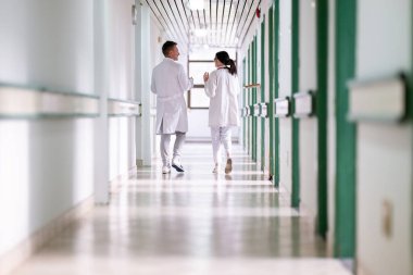 Beyaz önlüklü iki doktor, hastane koridorunda yürüyüp tıbbi belgeleri tartışıyorlar. Takım çalışmalarını ve profesyonel sağlık hizmetlerini ön plana çıkarıyorlar.. 