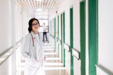 Kendine güvenen, steteskoplu genç bir bayan doktor. Parlak bir hastane koridorunda dururken gülümsüyor.. 