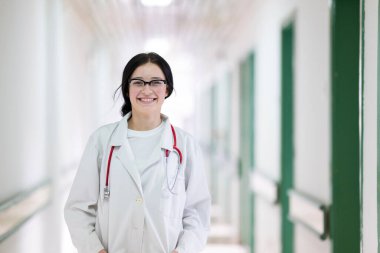 Kendine güvenen, steteskoplu genç bir bayan doktor. Parlak bir hastane koridorunda dururken gülümsüyor.. 