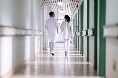 Beyaz önlüklü iki doktor, hastane koridorunda yürüyüp tıbbi belgeleri tartışıyorlar. Takım çalışmalarını ve profesyonel sağlık hizmetlerini ön plana çıkarıyorlar.. 