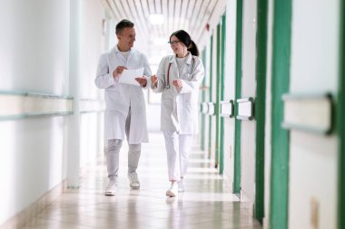 Beyaz önlüklü iki doktor, hastane koridorunda yürüyüp tıbbi belgeleri tartışıyorlar. Takım çalışmalarını ve profesyonel sağlık hizmetlerini ön plana çıkarıyorlar.. 
