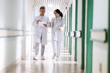 Beyaz önlüklü iki doktor, hastane koridorunda yürüyüp tıbbi belgeleri tartışıyorlar. Takım çalışmalarını ve profesyonel sağlık hizmetlerini ön plana çıkarıyorlar.. 