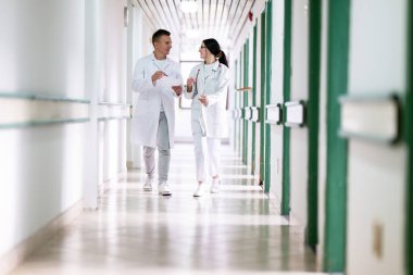 Beyaz önlüklü iki doktor, hastane koridorunda yürüyüp tıbbi belgeleri tartışıyorlar. Takım çalışmalarını ve profesyonel sağlık hizmetlerini ön plana çıkarıyorlar.. 