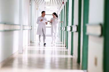Beyaz önlüklü iki doktor, hastane koridorunda yürüyüp tıbbi belgeleri tartışıyorlar. Takım çalışmalarını ve profesyonel sağlık hizmetlerini ön plana çıkarıyorlar.. 