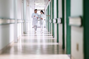 Beyaz önlüklü iki doktor, hastane koridorunda yürüyüp tıbbi belgeleri tartışıyorlar. Takım çalışmalarını ve profesyonel sağlık hizmetlerini ön plana çıkarıyorlar.. 