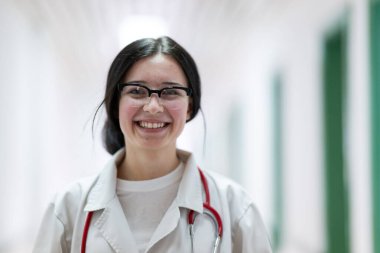 Kendine güvenen, steteskoplu genç bir bayan doktor. Parlak bir hastane koridorunda dururken gülümsüyor.. 
